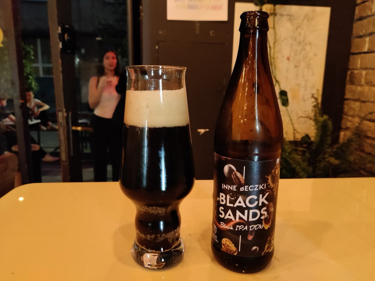 Ciemne piwo Black IPA nalane do kieliszka obok butelki na żółtym stoliku